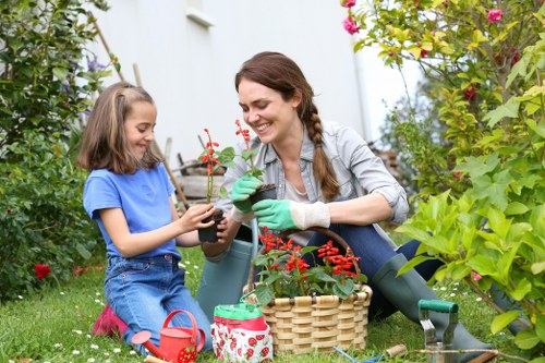 In a lush, well-maintained garden, a woman and a young girl are engaged in planting activities amidst vibrant greenery and flowering plants, including red and pink blossoms. The woman, with brown hair and wearing gardening gloves, holds a small potted plant, while the girl, dressed in a blue t-shirt and jeans, assists by holding the pot and smiling. They are seated on a grassy lawn with a variety of garden tools and supplies nearby, including a red watering can, a small bucket, and a wicker basket, indicating active gardening. The background features a white wall with climbing plants and shrubs, providing a peaceful outdoor setting typical of a residential garden in Acton, London. The scene captures a moment of outdoor activity under natural daylight, emphasizing garden care, planting, and gardening enjoyment, consistent with landscaping and lawn care services offered by Landscapers Acton.