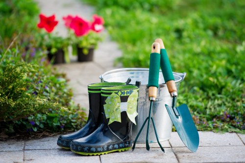 The image displays a tidy garden scene with a paved pathway bordered by lush green grass and various plants. In the foreground, a pair of dark rubber gardening boots with a green and black pattern rest on the paving, with a pair of gardening gloves partially tucked inside one of the boots. Nearby, a small metal bucket holds gardening hand tools, including a trowel with a blue handle and a hand fork with a wooden handle, both placed upright. In the background, vibrant pink and red flowering plants in black pots sit on the pathway, surrounded by dense greenery and shrubbery. The outdoor environment appears well-maintained and suitable for gardening activities, suggesting a landscaped garden area with a focus on practical outdoor maintenance, typical of services provided by Landscapers Acton in the Acton area, postcode W3, London. The weather looks bright and clear, with natural light illuminating the scene, emphasizing the fresh, natural tones of the garden and gardening tools, aligning with sustainable and eco-friendly gardening practices promoted on the company's Recycling and Sustainability page.