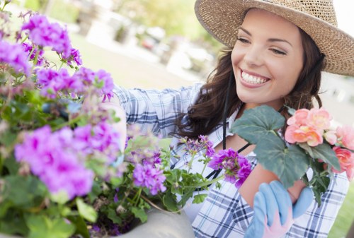 In the foreground of the image, a woman is engaged in gardening activities in an outdoor garden space, wearing a wide-brimmed straw hat, light blue gloves, and a plaid shirt. She is smiling while tending to a variety of flowering plants and shrubs, which include vibrant purple, pink, and peach blossoms. The garden features a neatly maintained lawn with lush green grass, bordered by flower beds and small shrubs, set in a spacious backyard environment with soft natural daylight. In the background, a paved pathway and a blurred view of trees and outdoor furniture are visible, indicating a well-kept landscaped yard suitable for outdoor activities. The scene reflects a pleasant, tranquil garden setting, highlighting horticultural care that a gardening service such as Landscapers Acton might provide, with an emphasis on flower bed maintenance, plant health, and overall lawn care in the Acton area near London, postcode W3.
