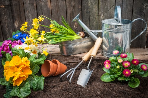 The image depicts a garden scene with freshly turned dark brown soil in the foreground, surrounded by various colourful potted flowers including yellow primroses, pink and purple primulas, and a cluster of blue and pink blossoms, all arranged on the ground. Several gardening tools are visible, such as a watering can, a trowel, a hand fork, and a small rake, positioned near the flower pots and soil, indicating ongoing planting or garden maintenance work. In the background, a weathered wooden fence provides a natural backdrop, suggesting an outdoor space in a residential garden, possibly in Acton or nearby. The overall scene reflects a well-organised gardening area suitable for planting new flowers or shrubs, with a focus on garden care and landscaping activities typical of professional garden maintenance services offered by companies like Landscapers Acton. The lighting is natural, indicating a clear, sunny day perfect for gardening activities, with natural tones of soil, vibrant flower colours, and weathered wood adding to the authentic gardening environment.