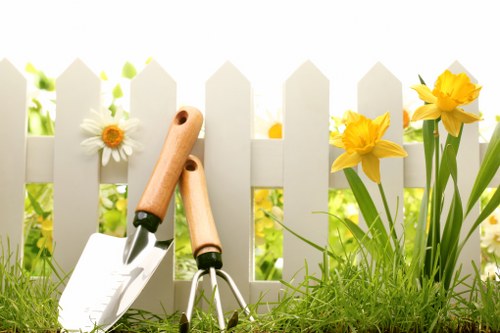 A close-up view of a small garden area featuring a white picket fence with pointed tops, behind which vibrant yellow daffodils and white daisies grow among lush green foliage. In the foreground, there is well-maintained grass with a dense, healthy appearance, and two gardening hand tools—a trowel and a hand cultivator with light wooden handles—are leaning against the fence, indicating recent or ongoing gardening activity. The scene is brightly lit with natural sunlight, suggesting a clear day, and the environment appears to be part of a front or backyard garden designed for decorative and outdoor leisure purposes. The arrangement emphasizes natural textures of the soil, plants, and wooden tools, consistent with gardening and lawn care services in the Acton area, aligning with professional landscaping and outdoor maintenance efforts by Landscapers Acton.