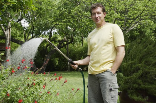 A man in a light yellow t-shirt and grey trousers stands in a well-maintained garden during daytime, holding a garden hose and watering a bed of red flowering plants. The garden features a lush, green lawn with dense shrubs and mature trees in the background, providing a shaded environment. Visible garden elements include a neatly edged flower bed, vibrant red flowers, and a backdrop of leafy hedges and tall trees, creating a natural and inviting outdoor space typical of landscaped gardens in Acton. The scene suggests outdoor maintenance activity, with the sunlight filtering through the foliage, highlighting the textures of grass, plants, and garden structures. This setting reflects the scope of professional gardening and planting services offered by Landscapers Acton, emphasizing lush greenery, flower displays, and garden care within a well-kept residential outdoor area in the Acton postcode area.