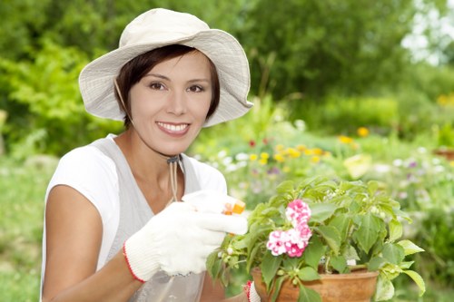 The image depicts a woman in a garden environment, wearing a white wide-brimmed hat and gardening gloves, smiling gently as she tends to a potted flowering plant with pink blooms. In the background, there is a landscaped outdoor space featuring well-maintained green lawns, a variety of shrubs and small trees, and flower beds with colorful plants, suggesting a healthy and cultivated garden. The ground surface appears to be a combination of soil, paving, and grass, with the garden bordered by natural greenery typical of a suburban or residential setting near Acton, London. The lighting indicates an outdoor scene during daylight, with natural sunlight illuminating the scene, emphasizing the vibrant greens, pinks, and natural tones of the garden elements. This image conveys professional gardening activities, suitable for outdoor lawn and plant maintenance services by Landscapers Acton, contributing to a tidy and thriving outdoor environment as part of comprehensive landscaping and garden care offerings in the local area.