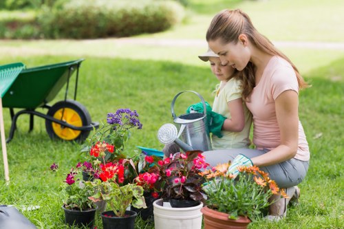 A woman and a young girl are engaged in gardening activities in a well-maintained outdoor garden space near Acton, with lush green grass and a backdrop of trees and shrubs. They are tending to a variety of potted flowering plants, including red, purple, and orange blossoms, placed on the grass. The woman is wearing a light pink T-shirt and grey jeans, while the girl is dressed in a yellow shirt. They are both wearing gardening gloves and focused on watering the plants, using a watering can. In the background, a green garden wheelbarrow with yellow wheels is visible, indicating outdoor landscaping effort. The scene reflects a neat, landscaped yard with a cultivated garden bed, a paved area or patio nearby, and a natural, sunny environment suitable for gardening and outdoor maintenance, demonstrating typical features landscaped by experts like Landscapers Acton.