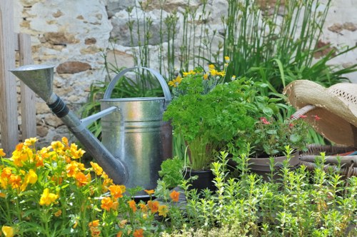 A small garden scene featuring a variety of plants and garden elements in front of a rustic stone wall. In the foreground, there is a dense patch of vibrant yellow and orange flowering plants, alongside green foliage with small leaves. Behind these, a silver metal watering can with a curved handle and a spout is placed among the plants, with a green potted shrub nearby. To the right, a partially visible straw hat with a decorative band rests on a garden surface, while surrounding plants include taller, grassy green plants and flowering bushes with pink blooms. The garden surface appears to be made of soil or mulch, and the overall lighting suggests a bright, partly sunny day, creating natural shadows and highlights. This outdoor garden space exemplifies typical gardening and landscaping elements that a professional service like Landscapers Acton might maintain or enhance, reflecting a well-kept, inviting outdoor environment in Acton, London, consistent with local gardening practices.