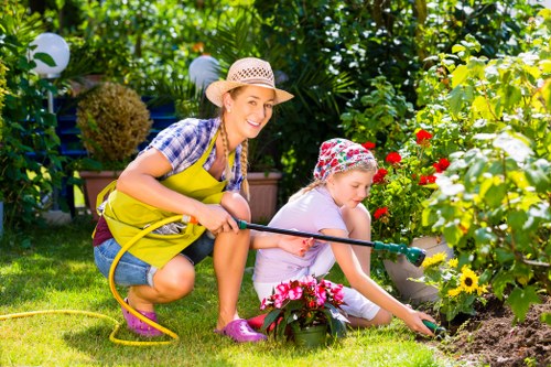 A woman and a young girl are gardening together in a lush backyard garden, with the woman wearing a wide-brimmed straw hat, a yellow apron, and casual clothing, smiling as she assists the girl. The girl is dressed in a pink headscarf and light-colored clothing, focused on planting or watering flowers. The garden features vibrant red, yellow, and pink flowers arranged in flower beds bordered by soil and a small grassy lawn with evenly cut, green grass. In the background, dense foliage, including bushes, trees, and a large shrub, provides privacy and a natural setting, while outdoor string lights hang among the greenery, suggesting a well-maintained outdoor space. The scene is brightly lit, indicating daytime with clear weather. The activity reflects typical outdoor gardening and maintenance practices promoted by local landscaping services such as Landscapers Acton, supporting the natural beauty and upkeep of gardens in the Acton area, postcode W3.