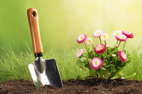 A garden scene features a partially visible wooden-handled trowel inserted into rich, dark soil in the foreground, indicating ongoing planting or soil preparation. To the right, a vibrant cluster of pink and white flowers with round, daisy-like blooms and green leafy foliage adds a splash of colour against the blurred green background, suggesting a well-maintained outdoor space typical of a suburban garden in Acton. The lawn surrounding the planting area displays healthy, even grass with a mix of light and dark green tones, and a neatly edged flower bed borders the planting zone, contributing to a tidy, organized appearance. Slight shadowing suggests natural daylight, possibly a clear or partly cloudy day, enhancing the natural colours of the plants and soil. This scene reflects professional gardening practices, suitable for garden maintenance, planting, and landscaping services provided by companies like Landscapers Acton, with an emphasis on soil preparation and ornamental plant care in a typical UK backyard setting.